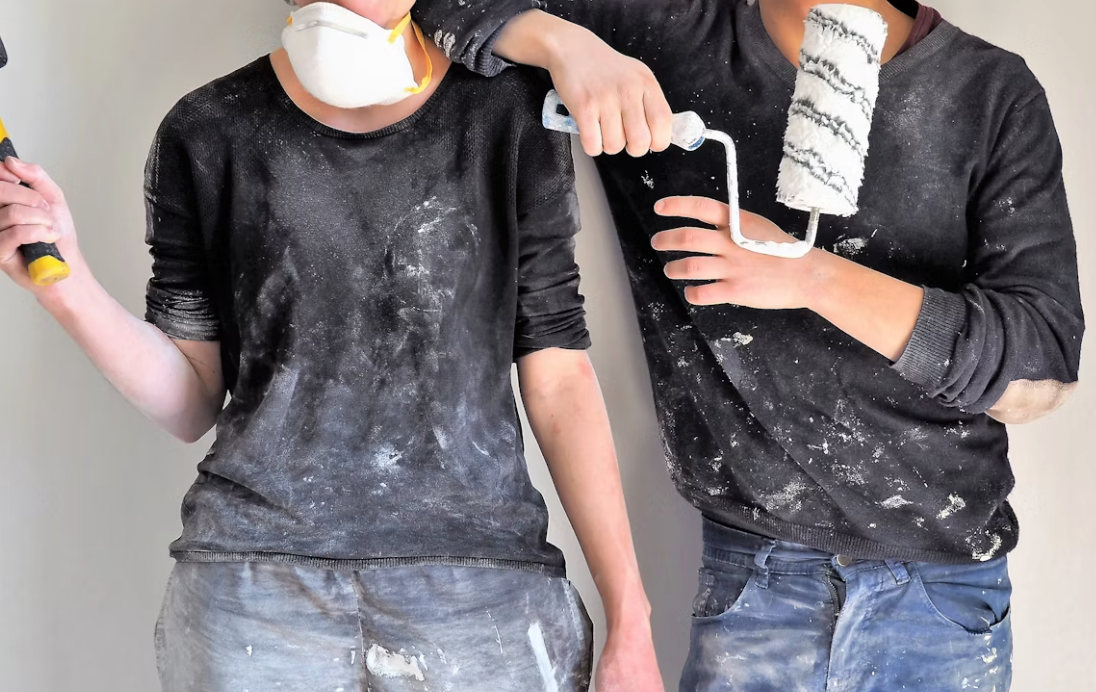 Two renovators in dust-covered work clothes holding a hammer and paint roller.