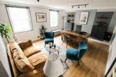 Home office with a wooden desk, teal velvet chairs, tan sofa, two windows.